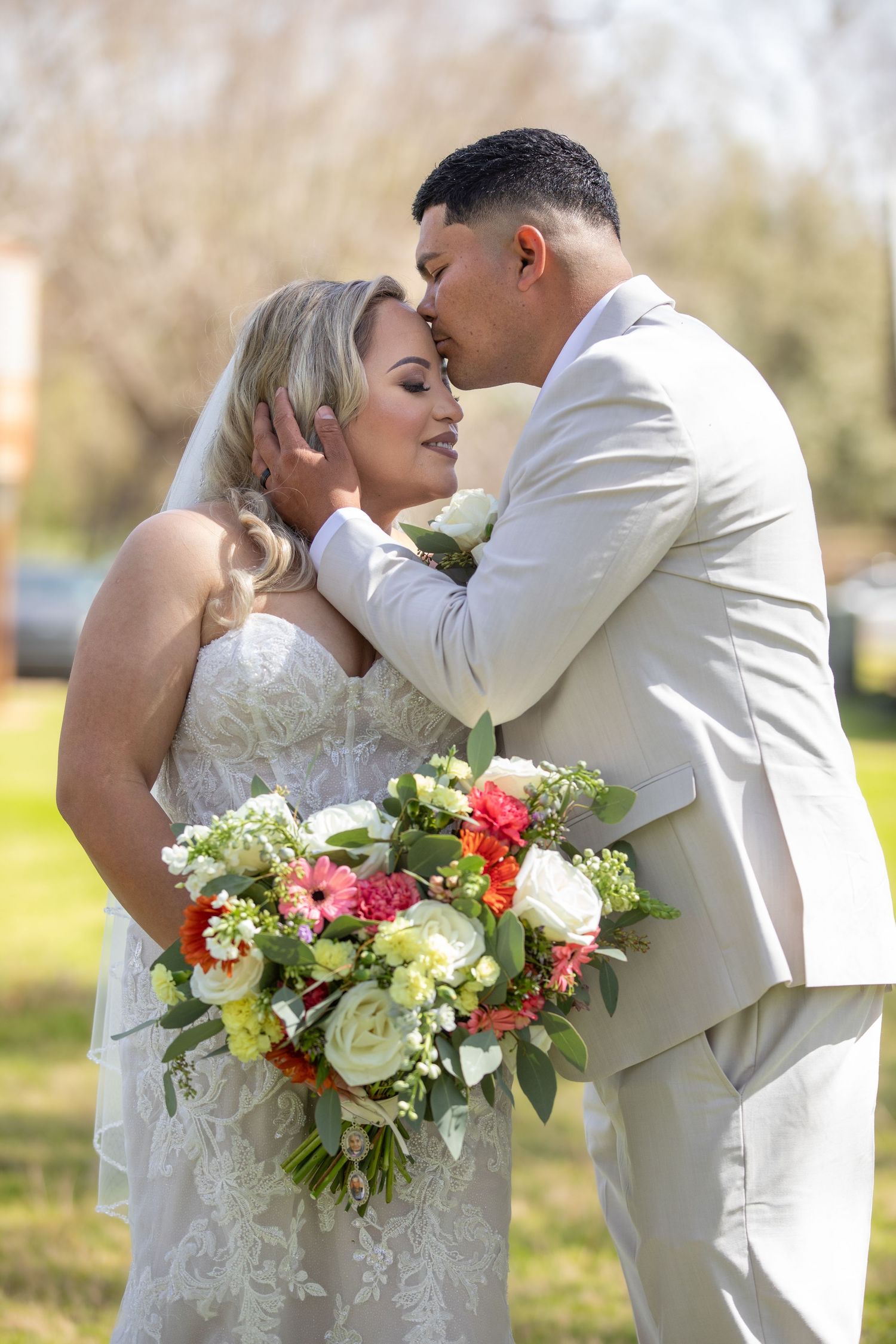 Groom in beige suit kisses bride in lace wedding dress while she holds a colorful bouquet outdoors.