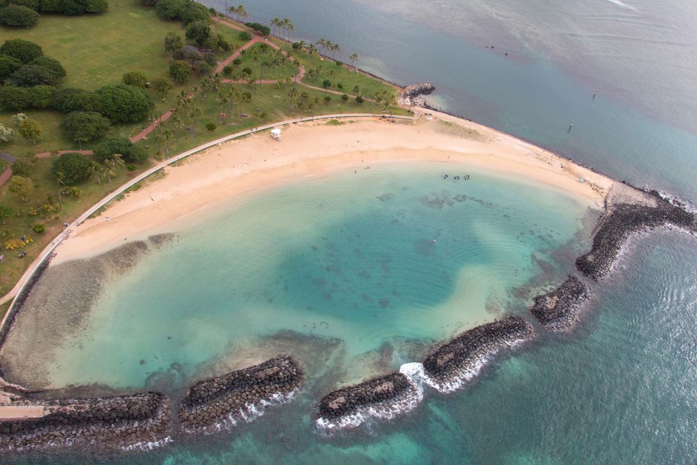 Aerial view of a curved beach with turquoise water and black volcanic rocks forming a natural breakwater.