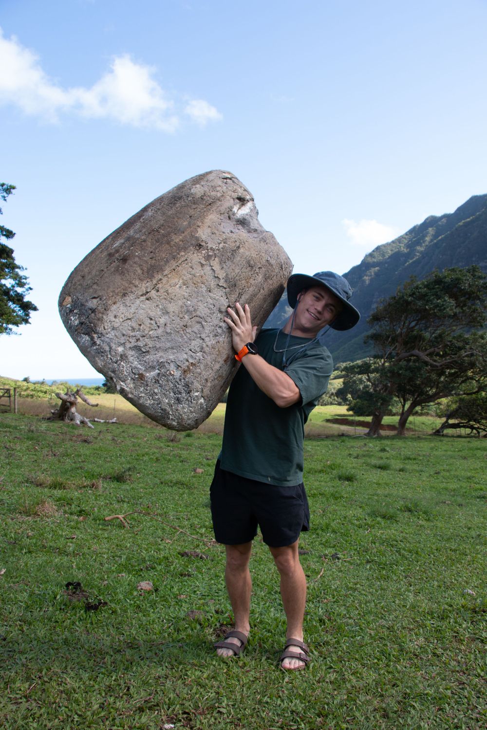 Hiker in dark clothing lifts massive boulder while standing in a grassy field with mountains in the background.