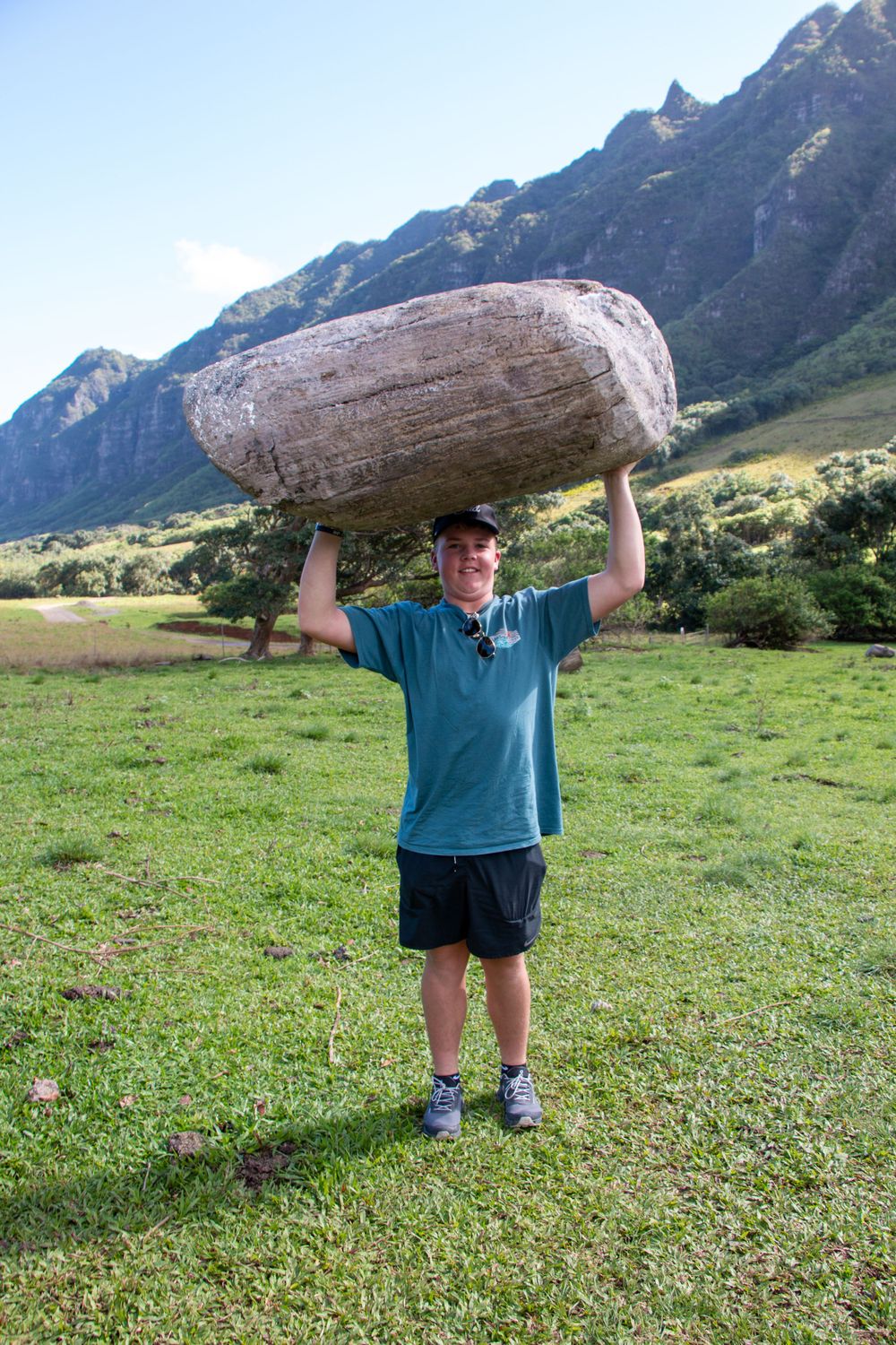 Someone lifts a large boulder overhead while standing in a grassy meadow with mountain peaks in the background.