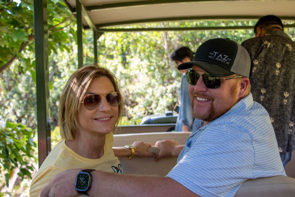 Two people enjoy a sunny outdoor meal under a covered area wearing sunglasses and casual attire.