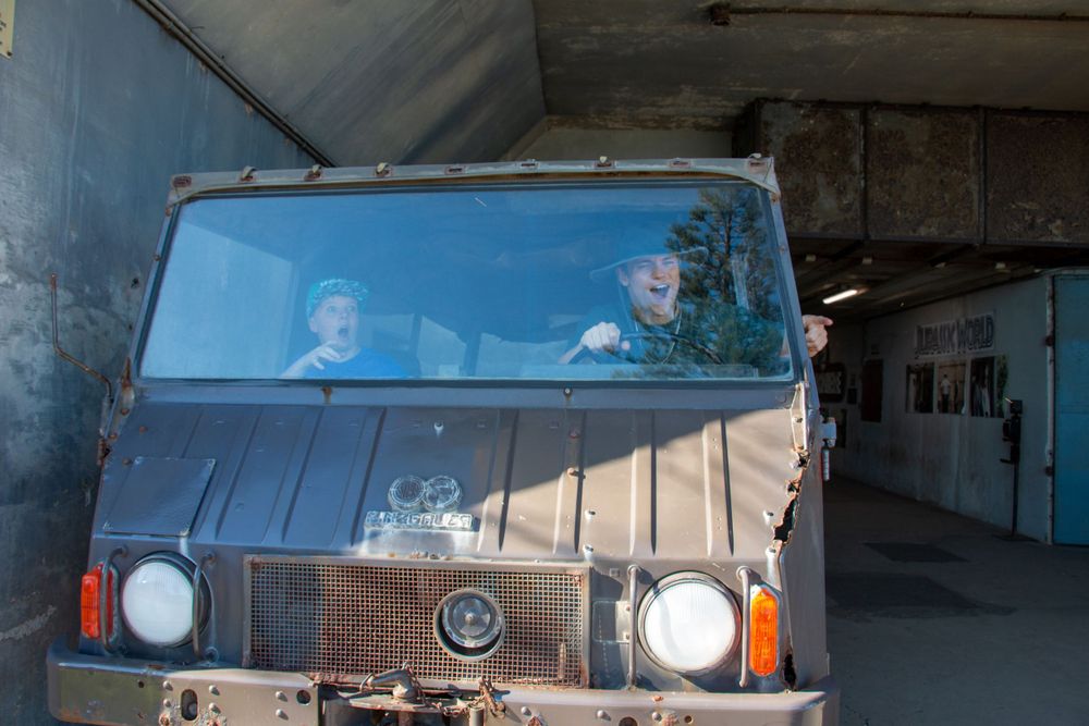 A vintage military truck parked under a covered area with two occupants visible through the windshield.