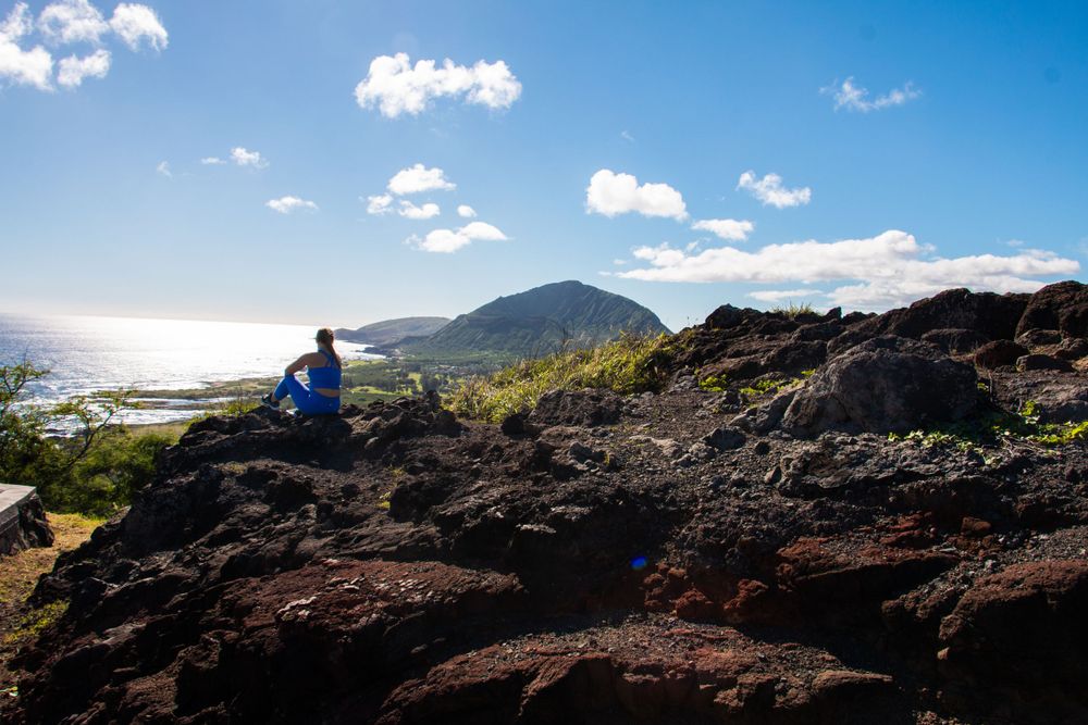 Figure sitting on volcanic rocks overlooking coastal mountains and ocean on a sunny day with white clouds.