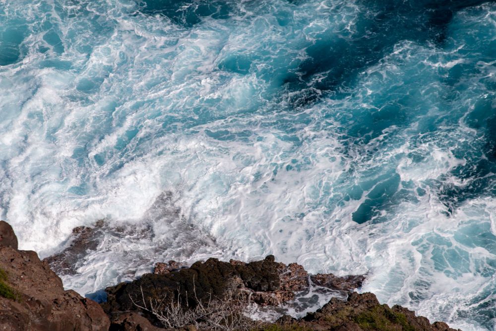 Aerial view of turbulent turquoise ocean waves crashing against rocky coastal cliffs.