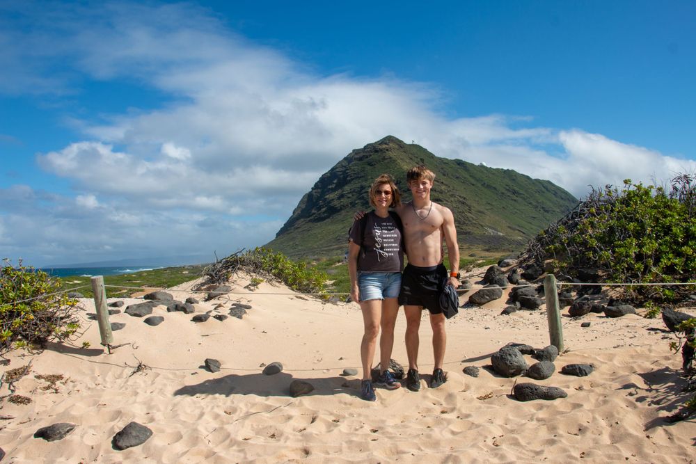 Two hikers standing on sandy trail with green mountain peak in background under blue cloudy sky.