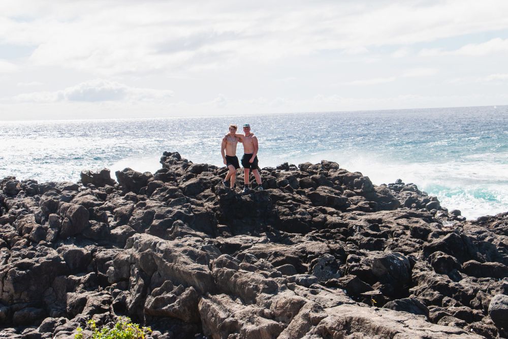 Two people stand on dark volcanic rocks as waves crash against the Hawaiian shoreline.