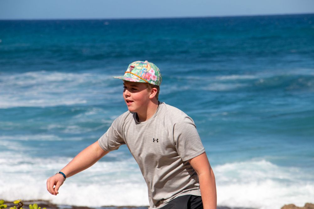 Person in a floral cap and gray t-shirt stands against a backdrop of bright turquoise ocean waves.