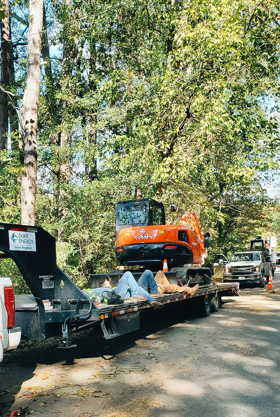 Orange excavator being transported on a flatbed trailer through a tree-lined road.