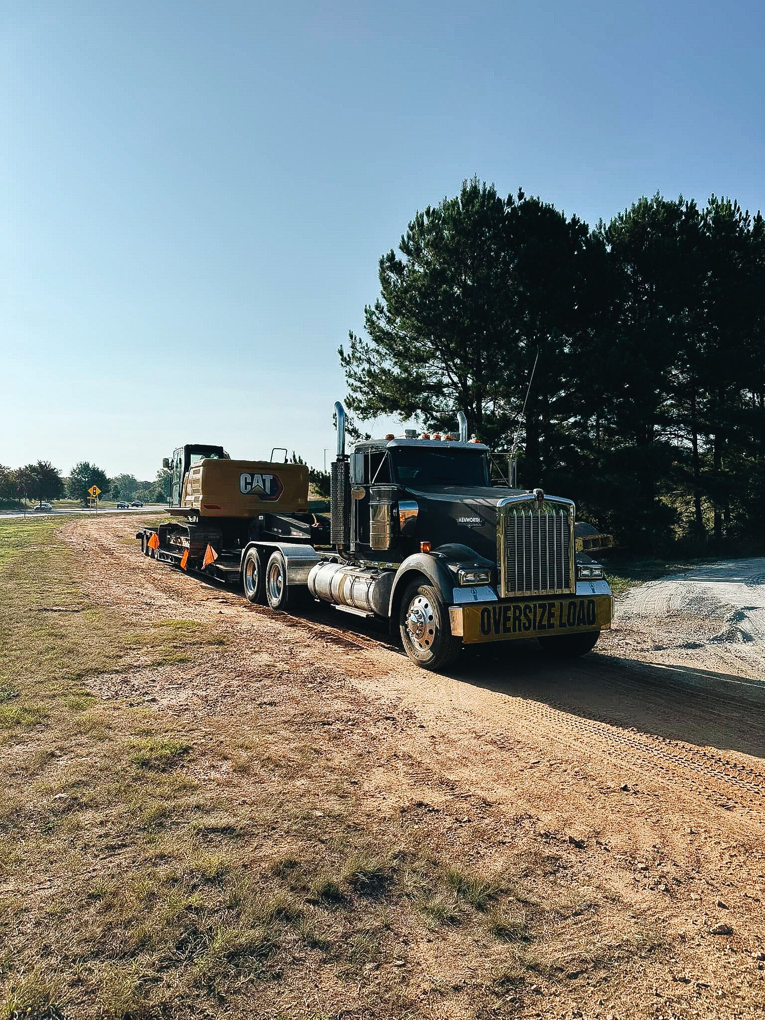 Semi truck hauling heavy equipment on dirt road near pine trees.