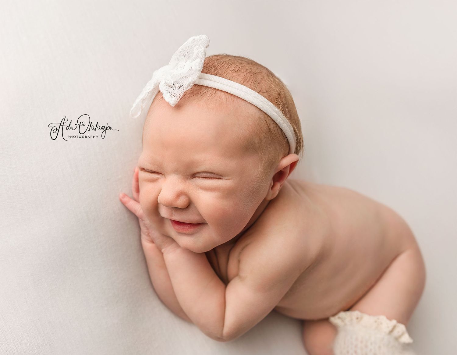 Newborn baby with white bow headband smiles while lying on side during professional photo session.