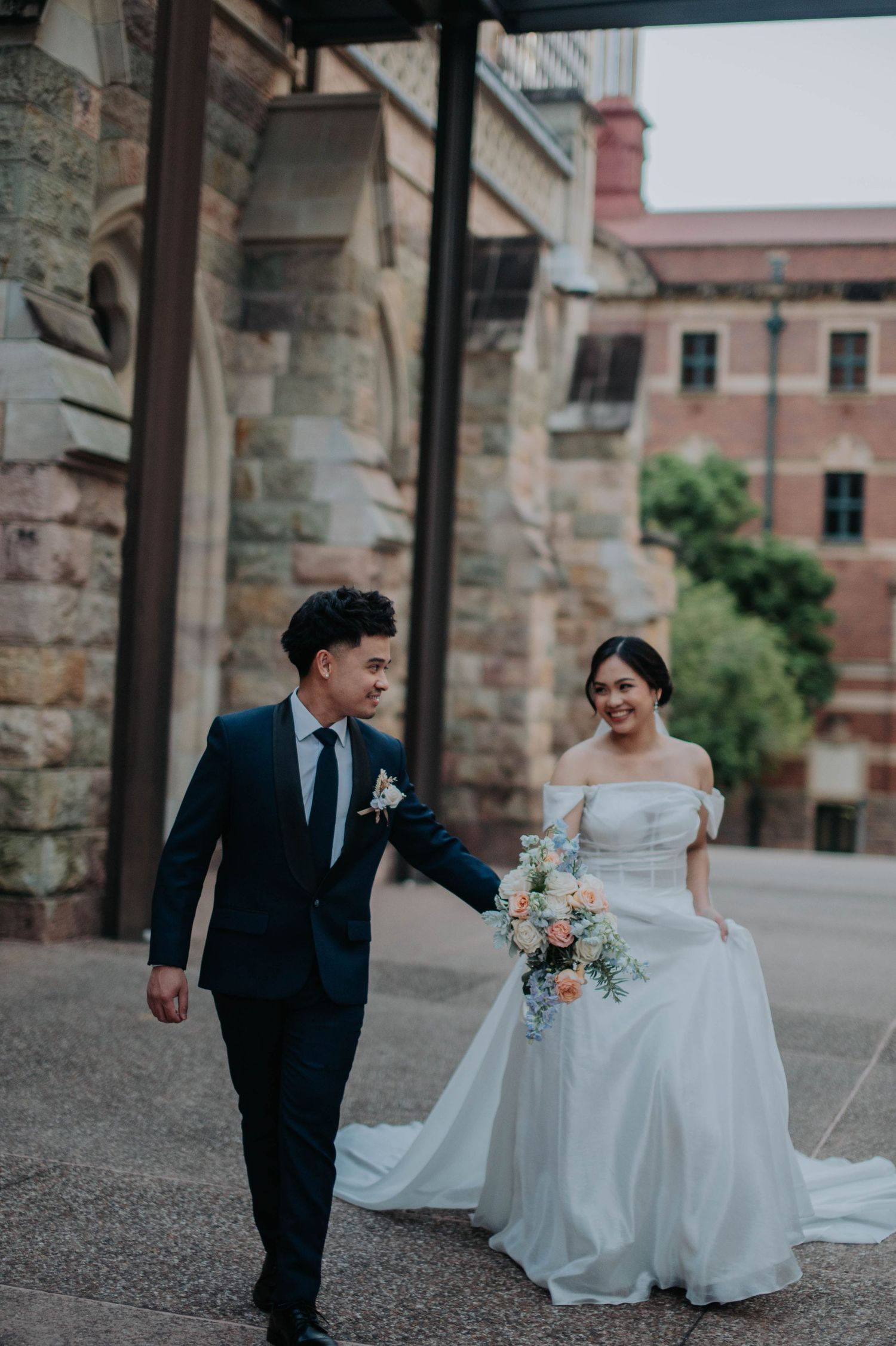 A couple in wedding attire walks together along a stone building with a trailing white gown.
