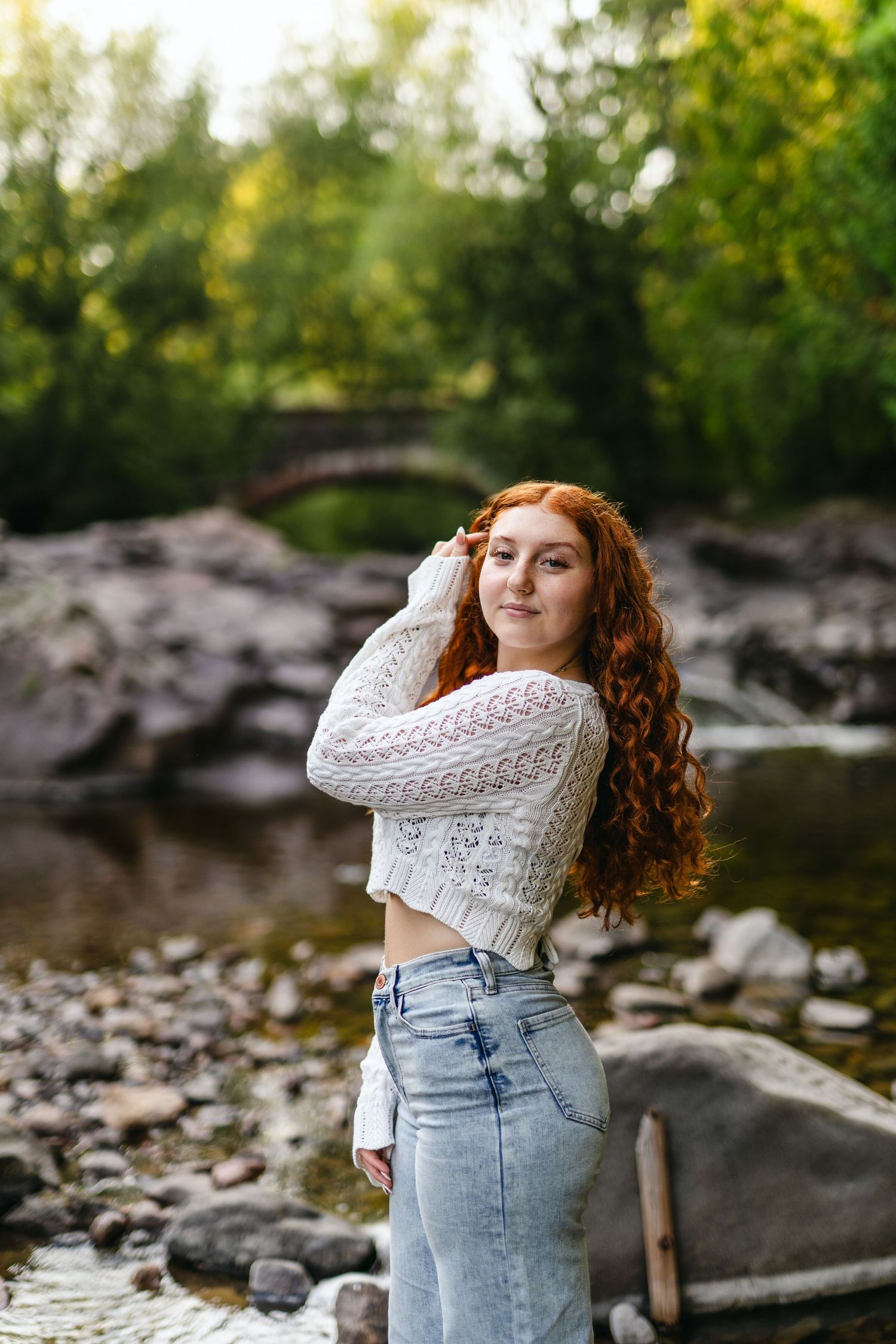 A person in a white knit sweater and jeans poses outdoors by rocky water in a natural setting.