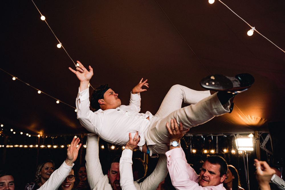 Celebratory moment at a wedding reception as guests lift someone in a white suit at the Lightfoot & Wolfville wedding venue.