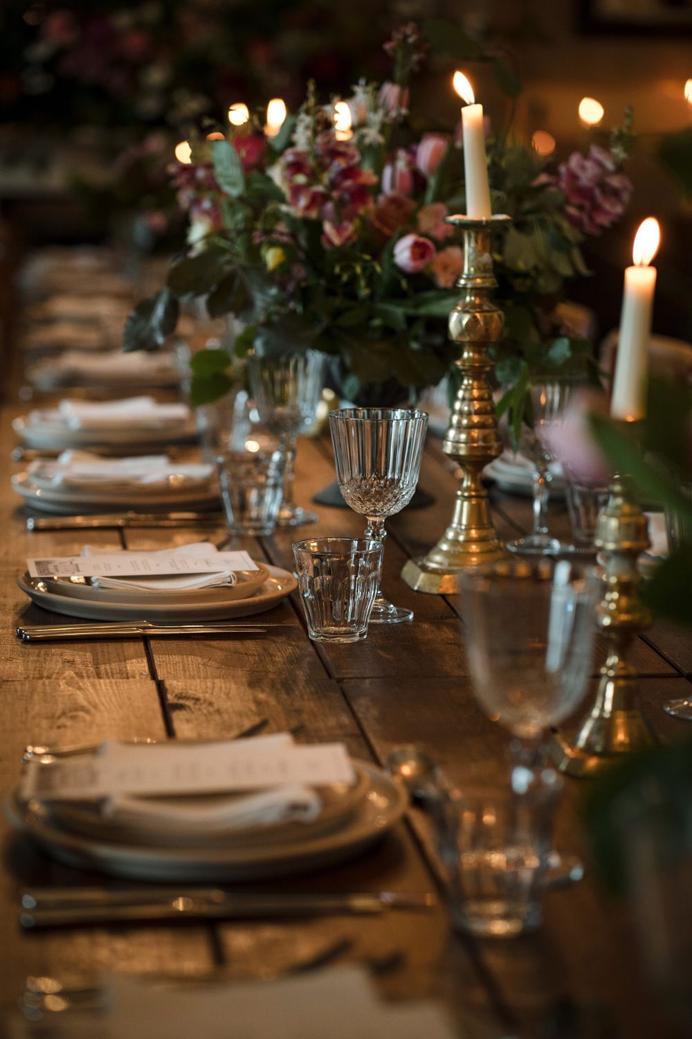 Elegant table setting with lit candles, crystal glasses and floral centerpiece on rustic wooden table in warm lighting.