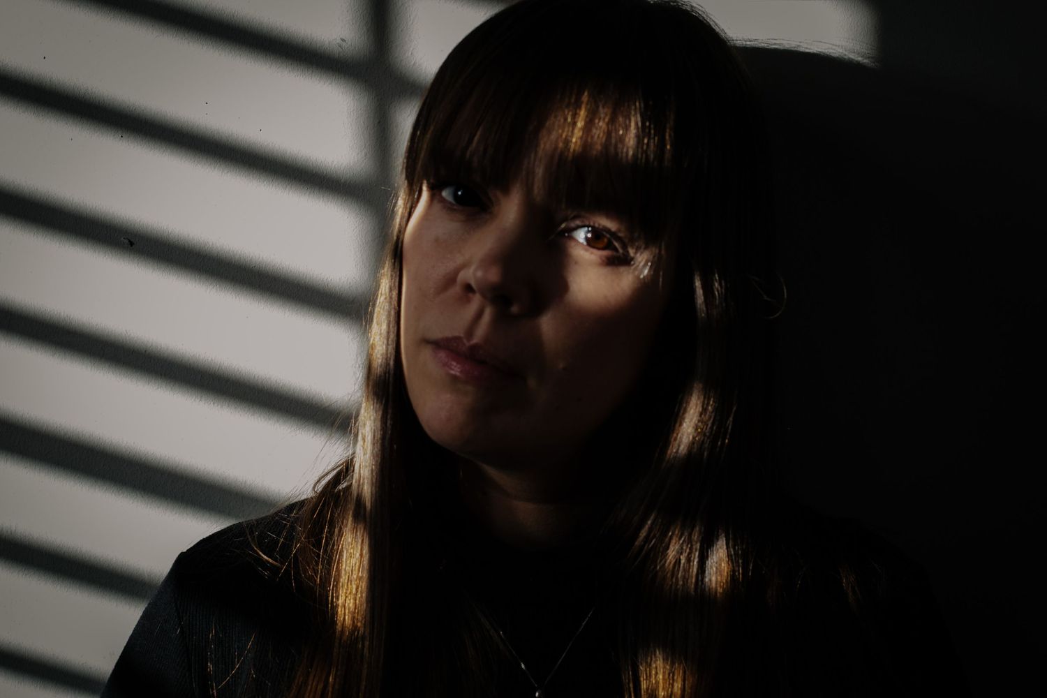 A dramatic portrait with light and shadow cast from window blinds illuminates someone in darkness with long bangs.