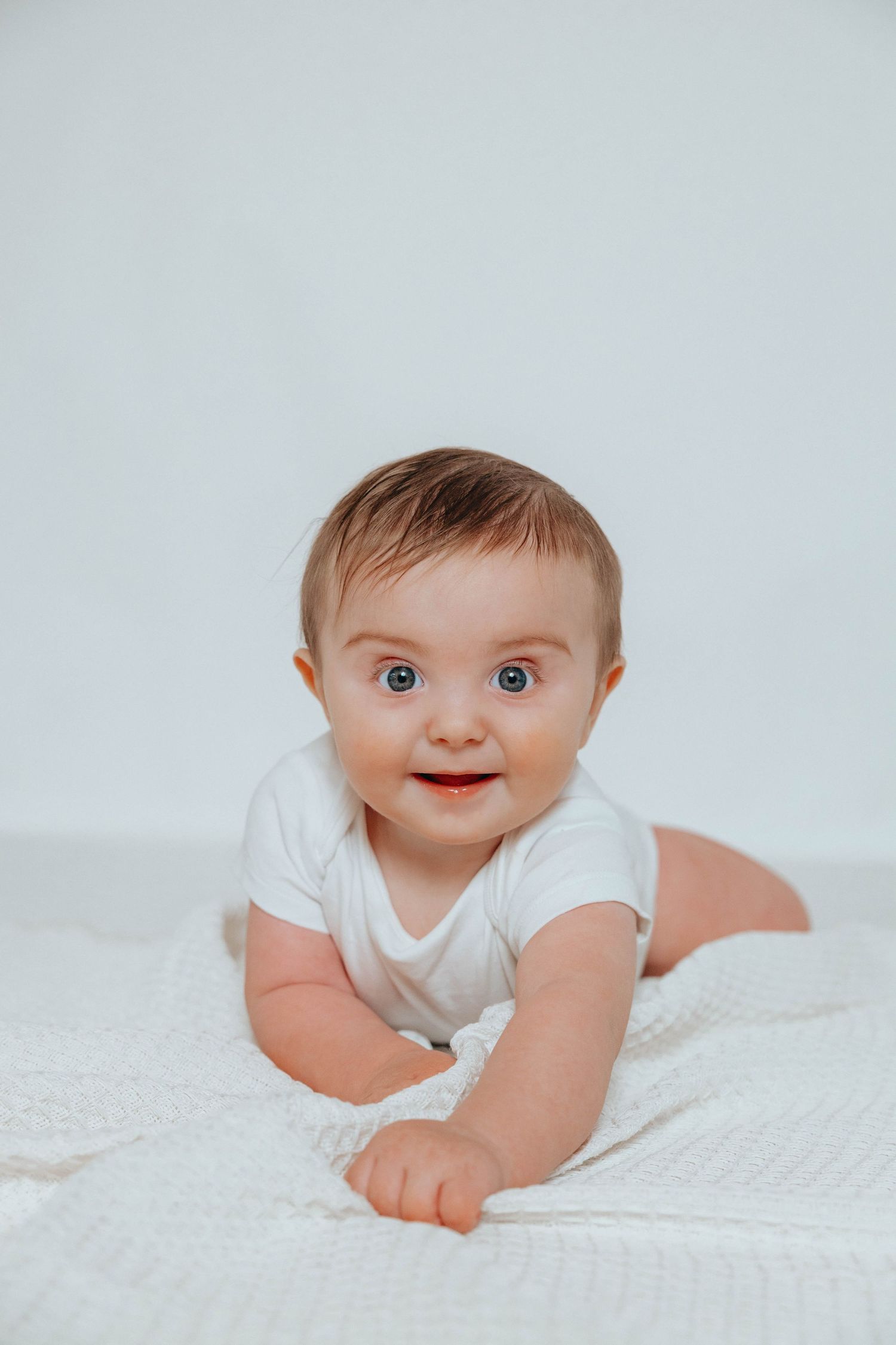Baby in white onesie lying on stomach on white blanket with bright expression and wide eyes.