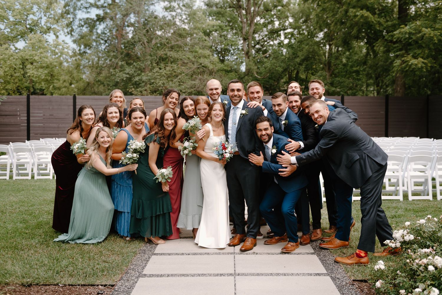 Wedding party poses together outdoors on stone path with white chairs and green trees in background.