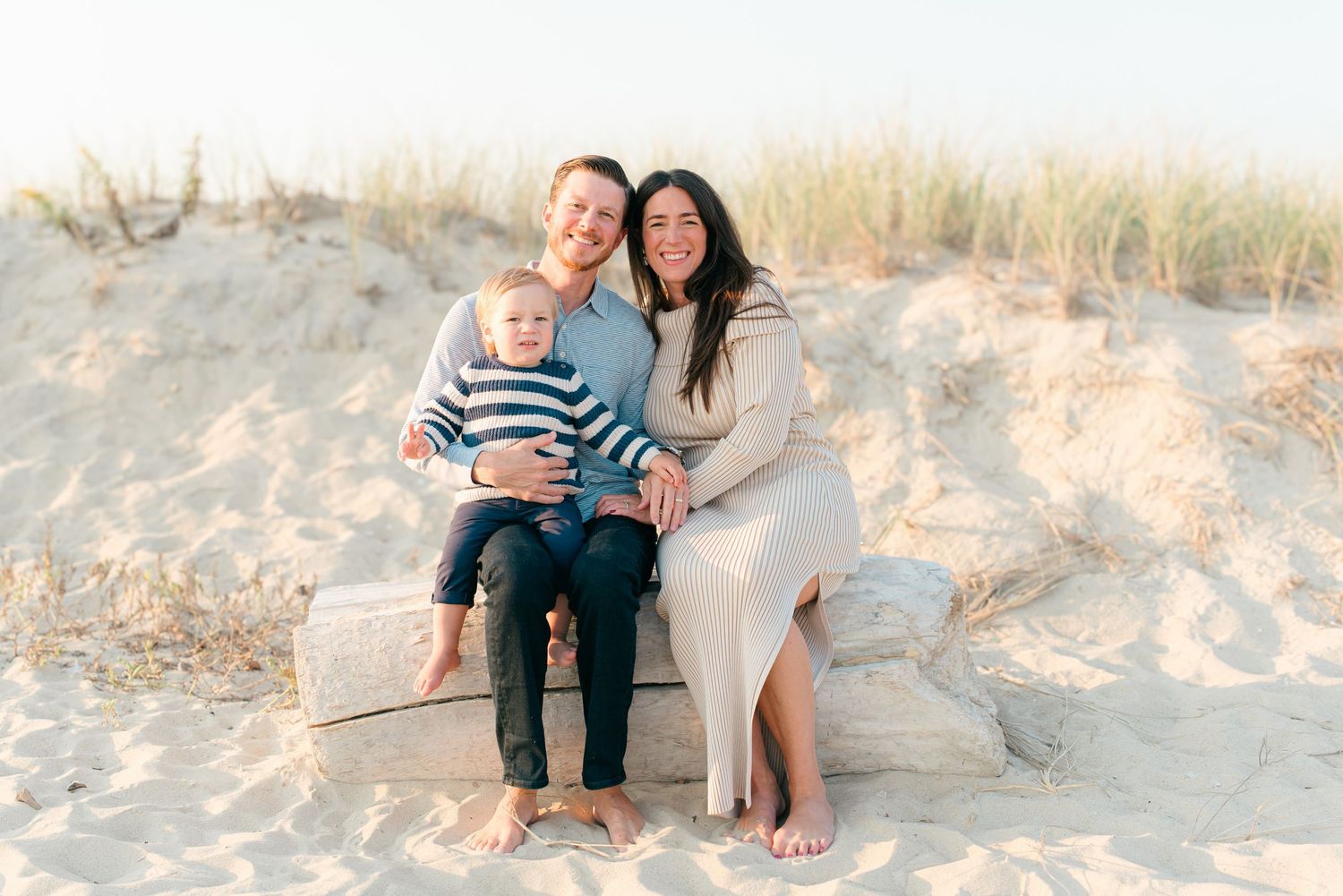 Family sitting together on a washed-up log at Spring Lake beach, smiling as the breeze lifts their hair and the fall sun glows behind them.