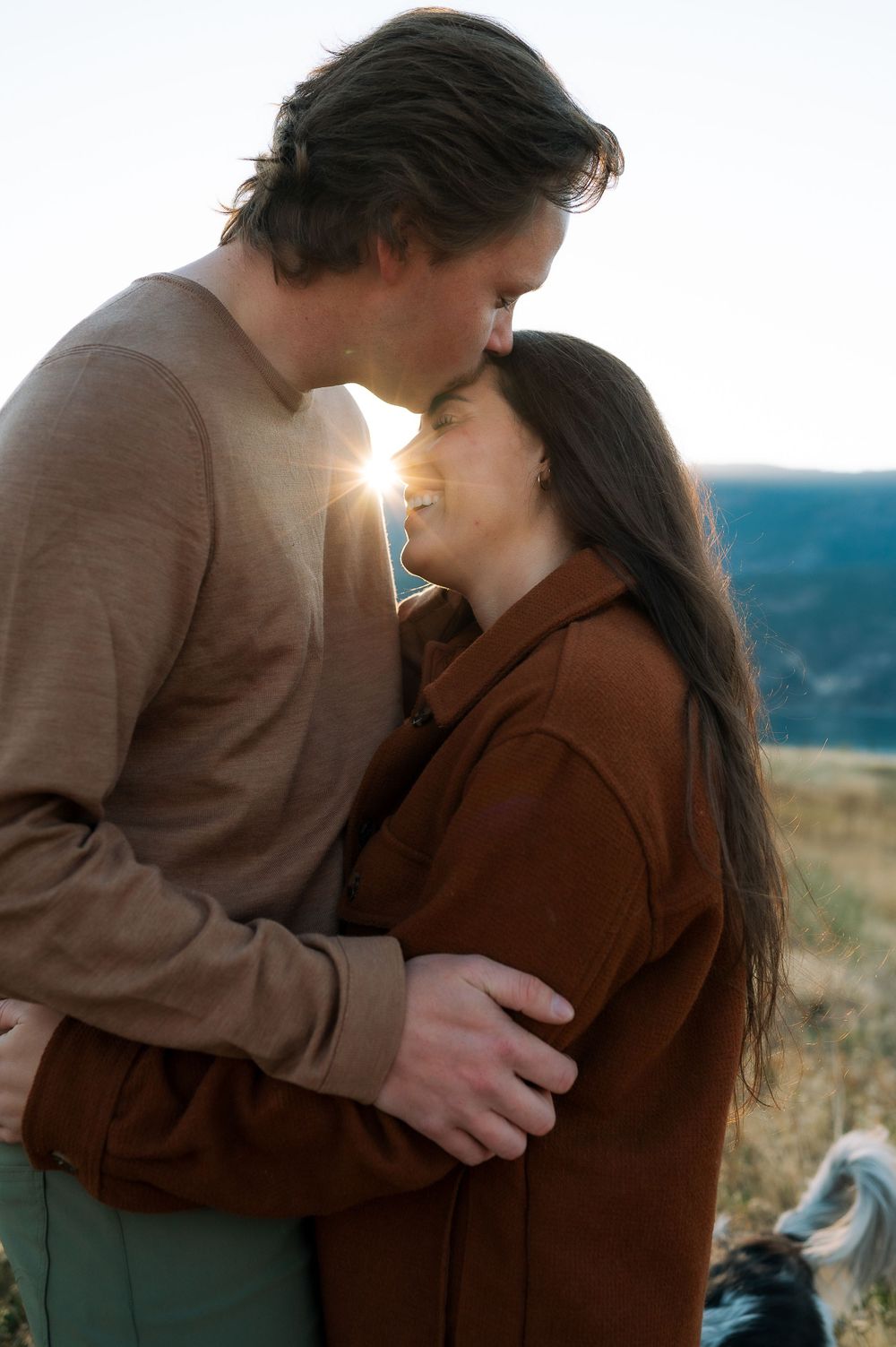Romantic couple embraces at sunset with sun flare peeking through in mountain landscape setting.