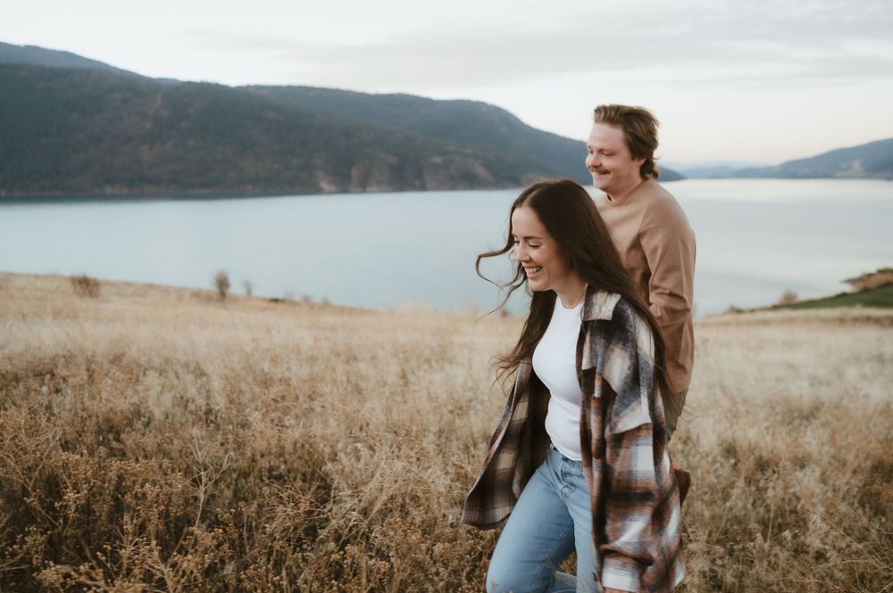 A couple shares a joyful moment on a hilltop overlooking a serene lake with mountains in the background.