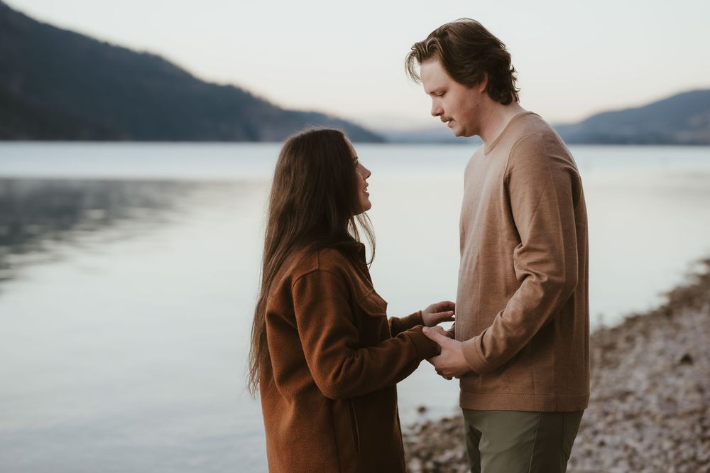 A tender moment between a couple holding hands by a tranquil lake surrounded by mountains at dusk.