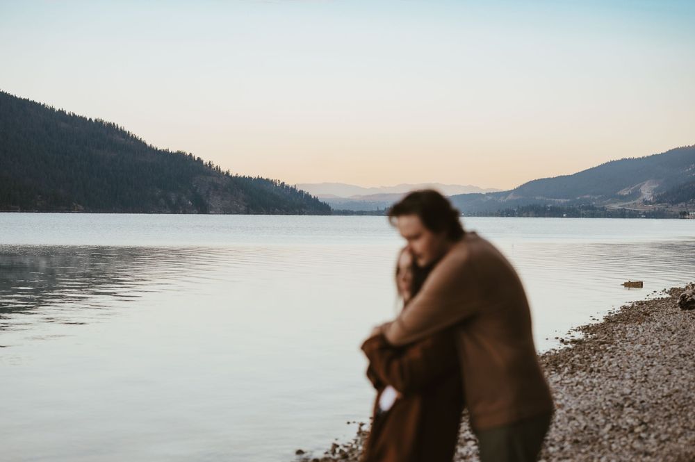 Couple in warm embrace by lakeside during golden hour with mountains in distance.