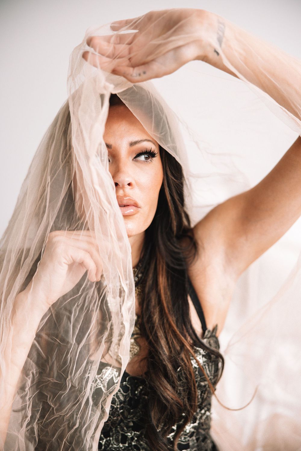 Alluring woman with long wavy dark hair holding a sheer blush veil above her face in a bright studio.
