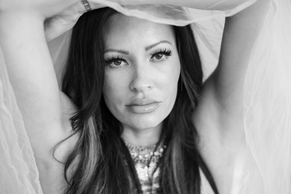 Black and white portrait of a beautiful woman with long dark hair holding sheer fabric above her head.