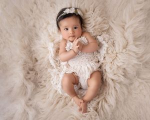 Baby in lace dress lying on circular cream fur rug during professional photoshoot.