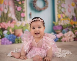 A baby in a pink ruffled dress sits on a blanket against a floral backdrop with a turquoise door and wreath.