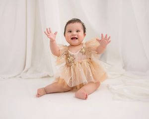 Baby wearing sparkly gold tulle dress sitting on white fabric backdrop with hands raised excitedly.
