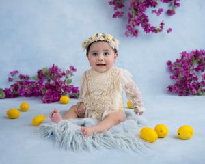 Baby with flower crown seated among yellow lemons and purple flowers against blue backdrop.