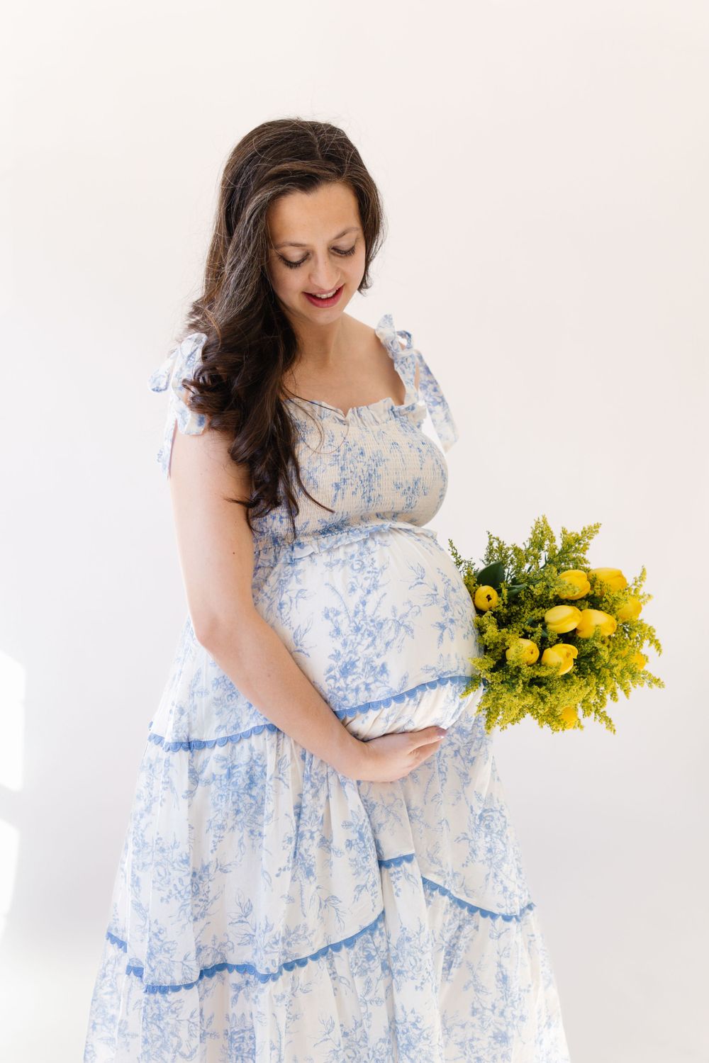 A person in a blue and white floral maternity dress holds a bright yellow spring bouquet against a light background.