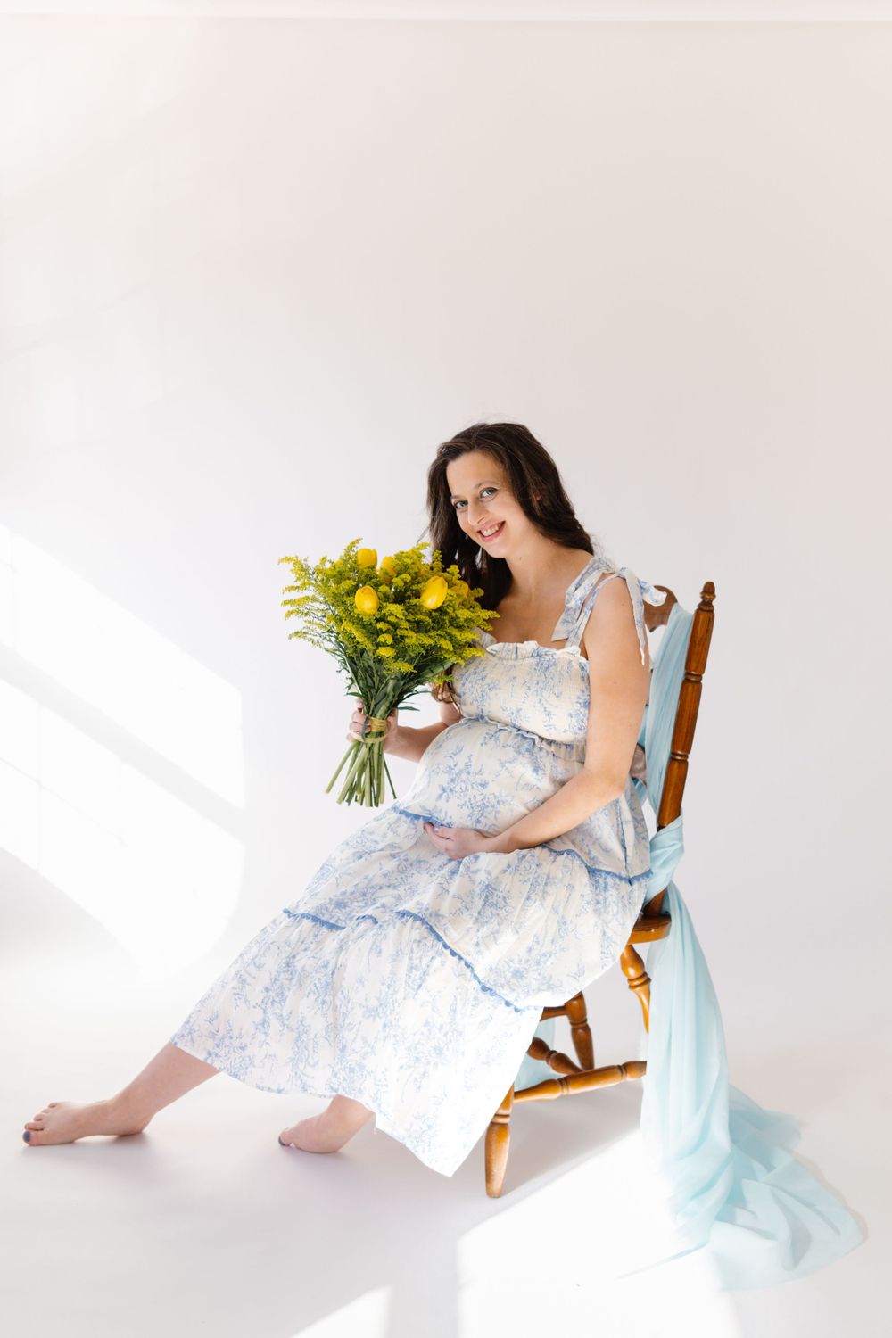 Expectant person in a flowy blue floral dress sits on wooden chair holding bright yellow flowers in a bright white studio.