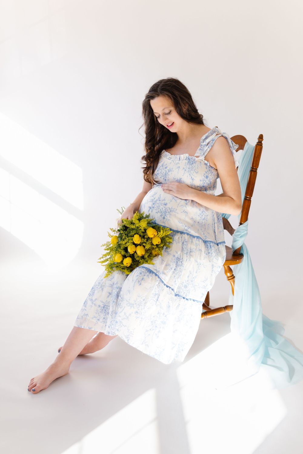 Person in flowing floral dress sits on wooden chair holding bright yellow wildflower bouquet in sunlit white room.