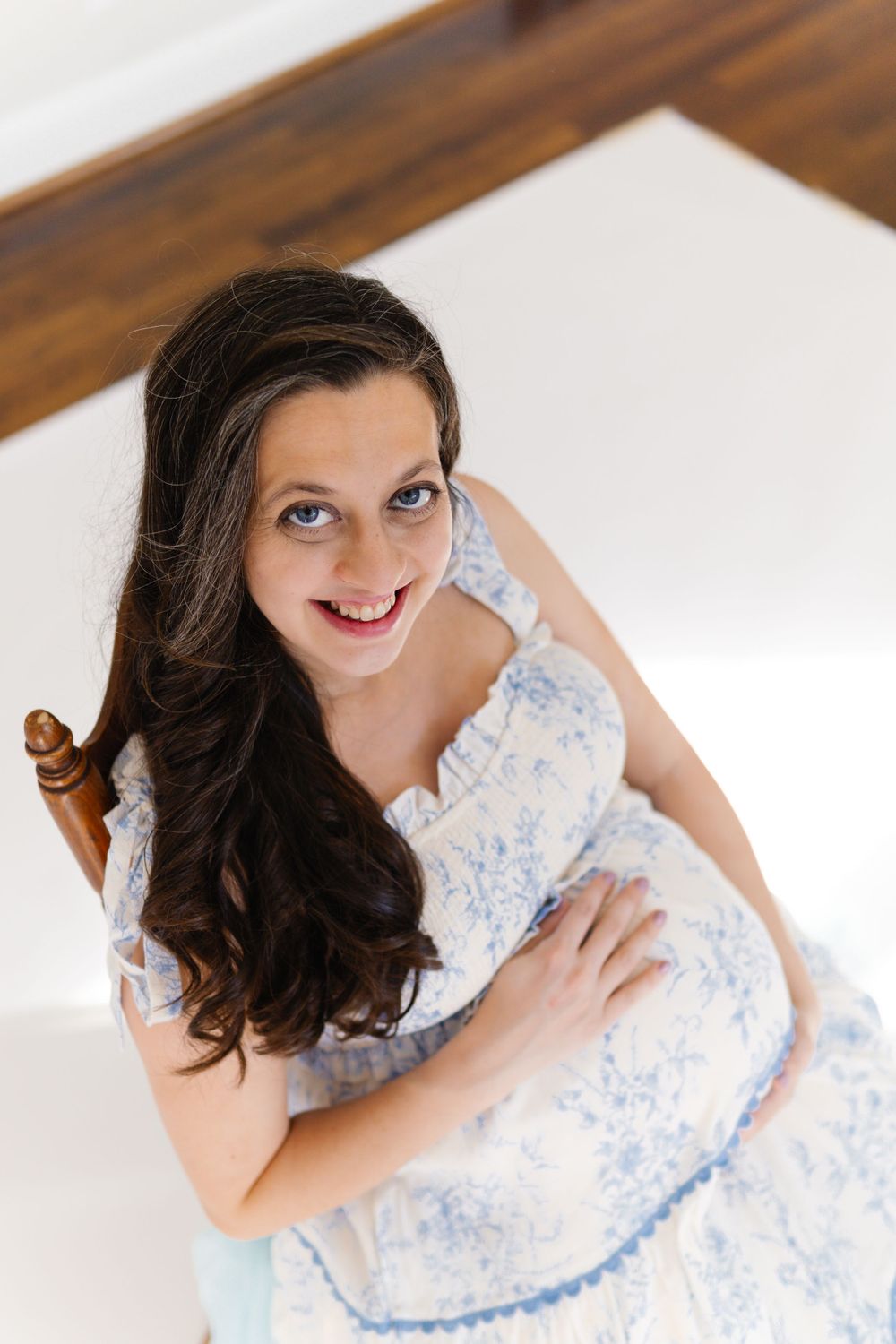 A woman in a blue and white floral dress poses while sitting on a wooden chair in a light-filled room.