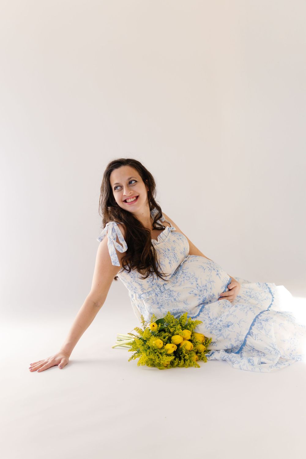 A maternity photo with yellow tulips features a woman in a delicate blue and white patterned dress.
