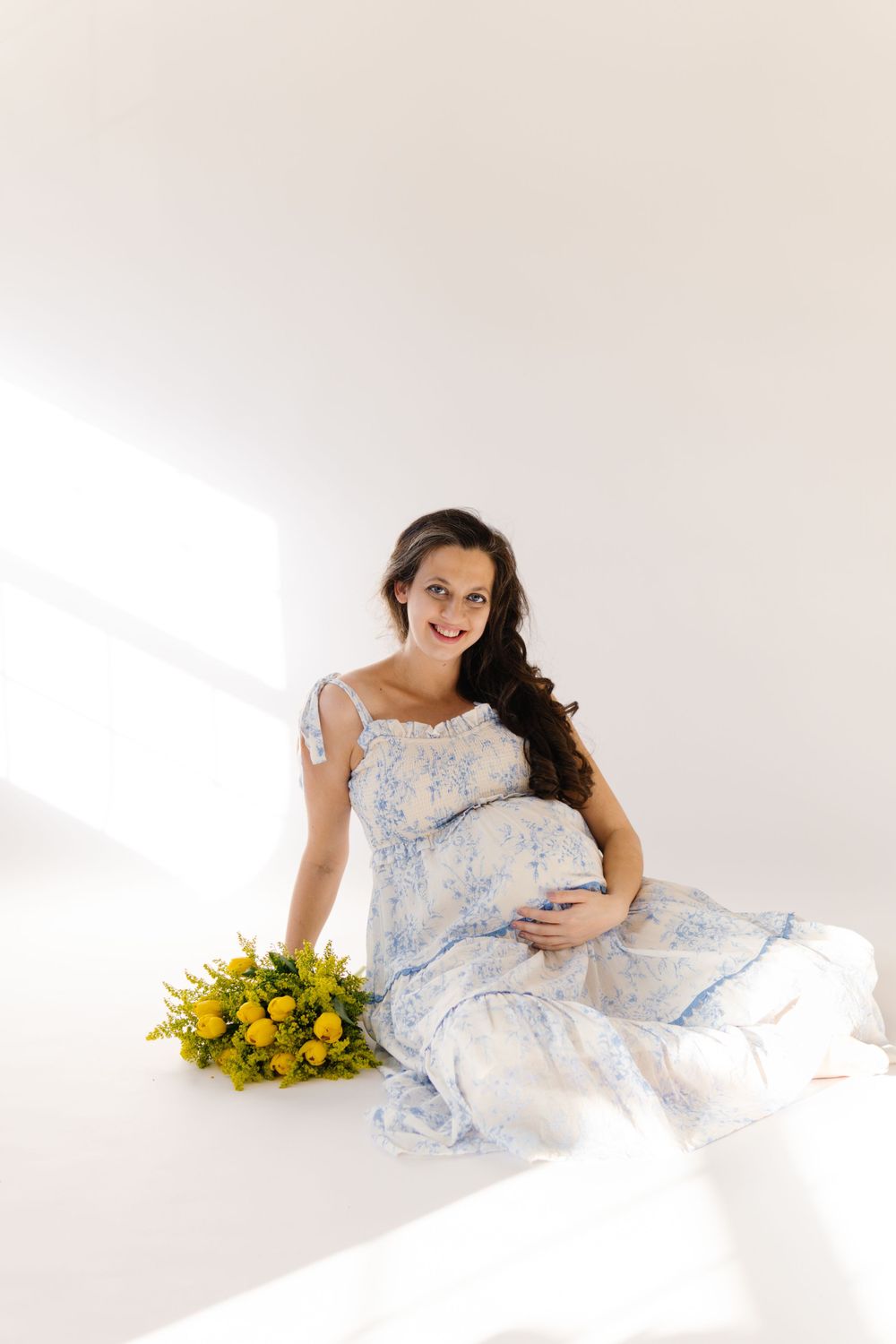 A pregnant woman in a flowing blue floral dress sits with a bouquet of yellow flowers on a white floor.