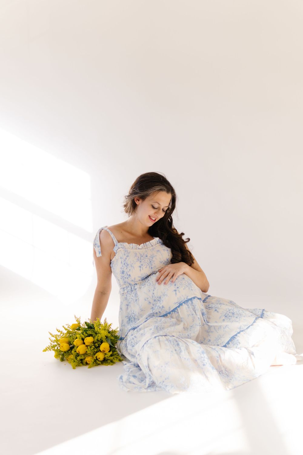 Woman in a flowing light blue dress sits gracefully with a bouquet of bright yellow flowers in a sunlit white room.