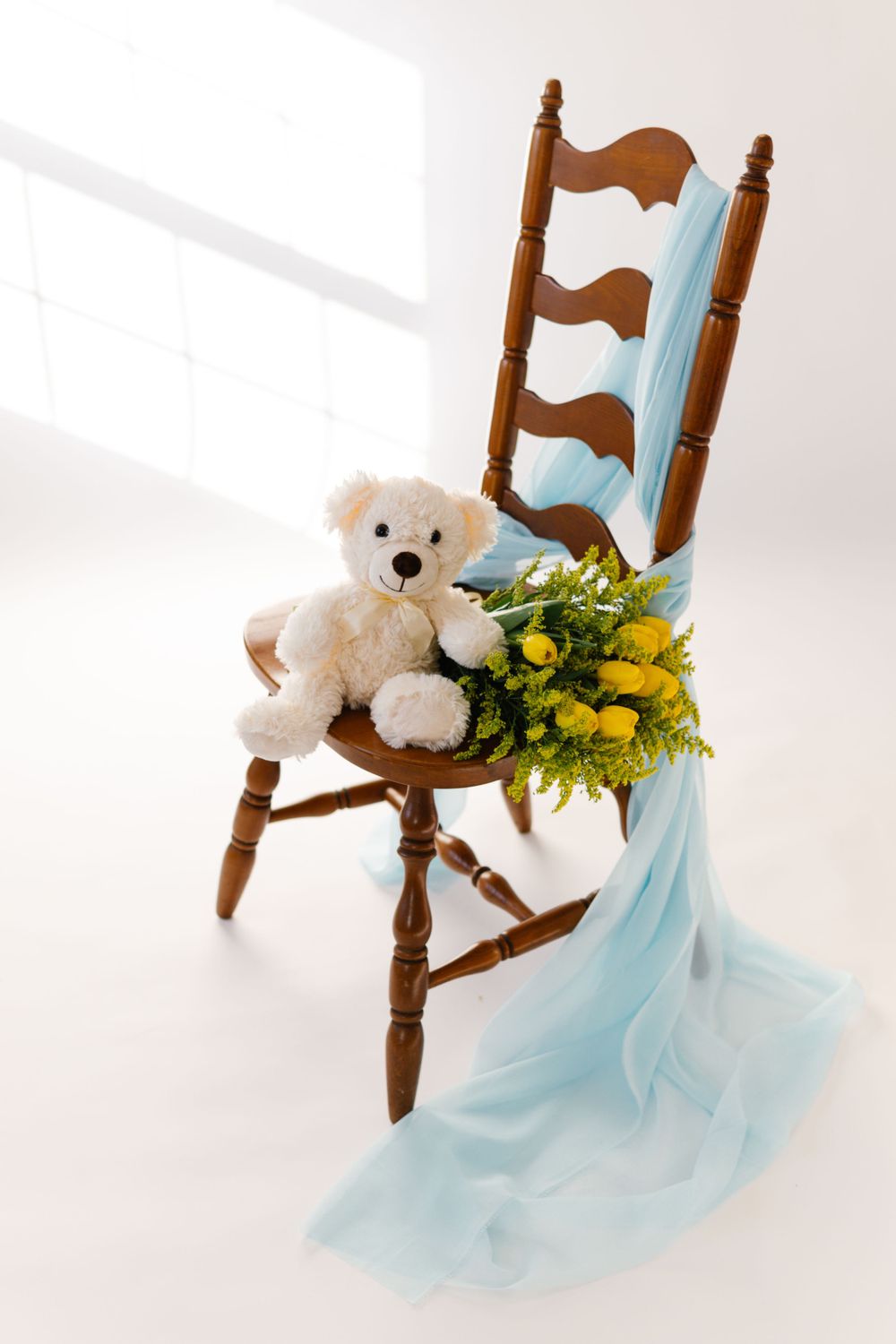 A vintage wooden chair decorated with light blue fabric, yellow flowers and a white teddy bear in a bright setting.