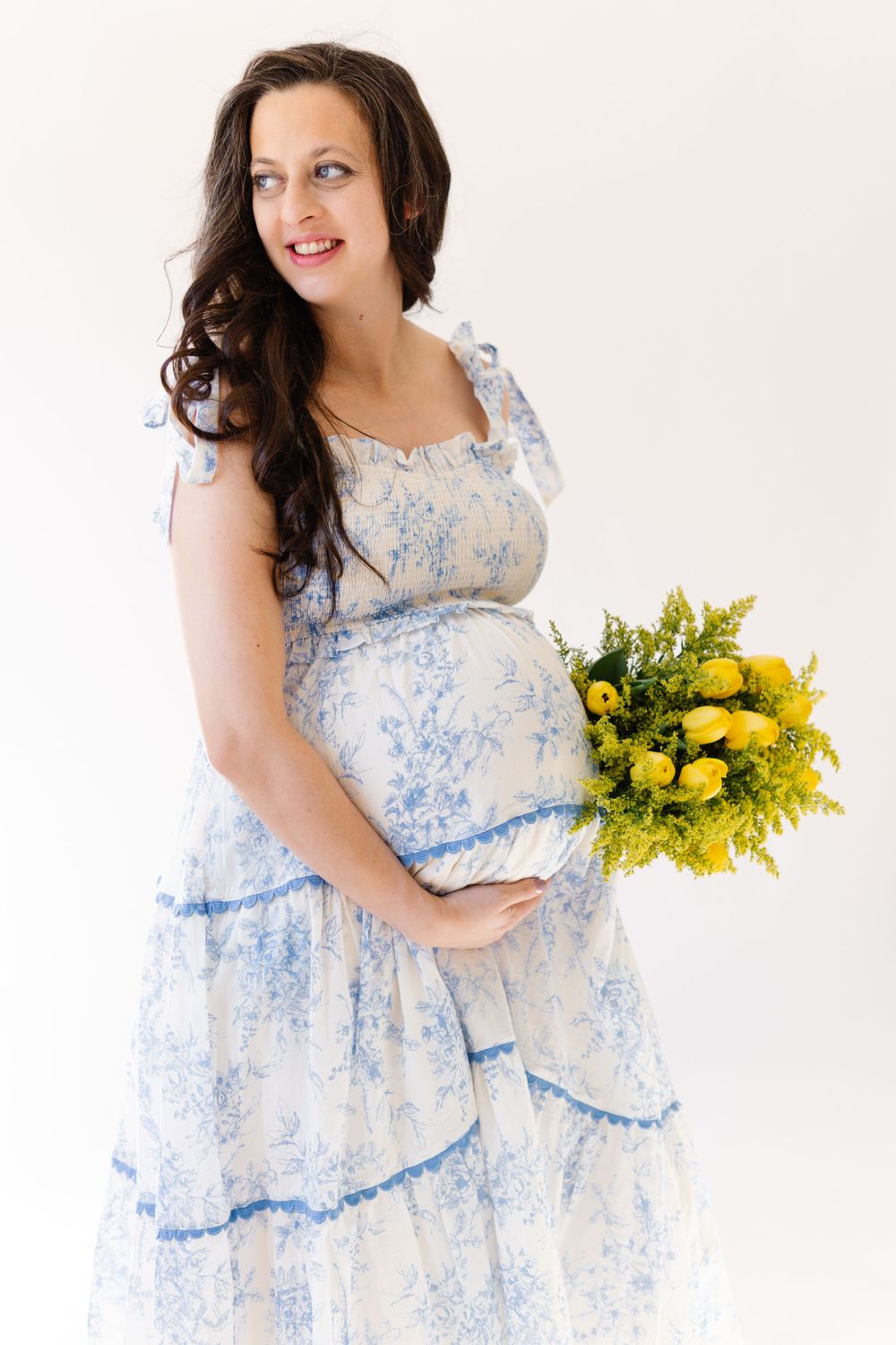 Expectant mother wearing delicate blue and white dress holds cheerful bouquet of yellow flowers.