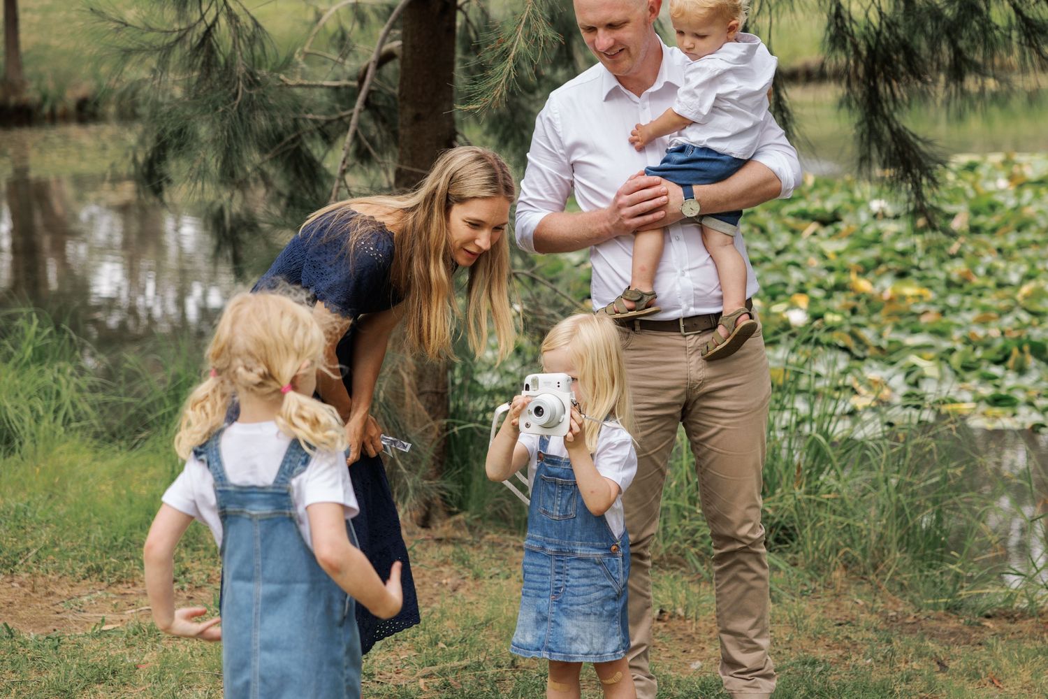 A family enjoys a casual outdoor gathering by a pond with their children and labrador retriever in a lush garden setting.