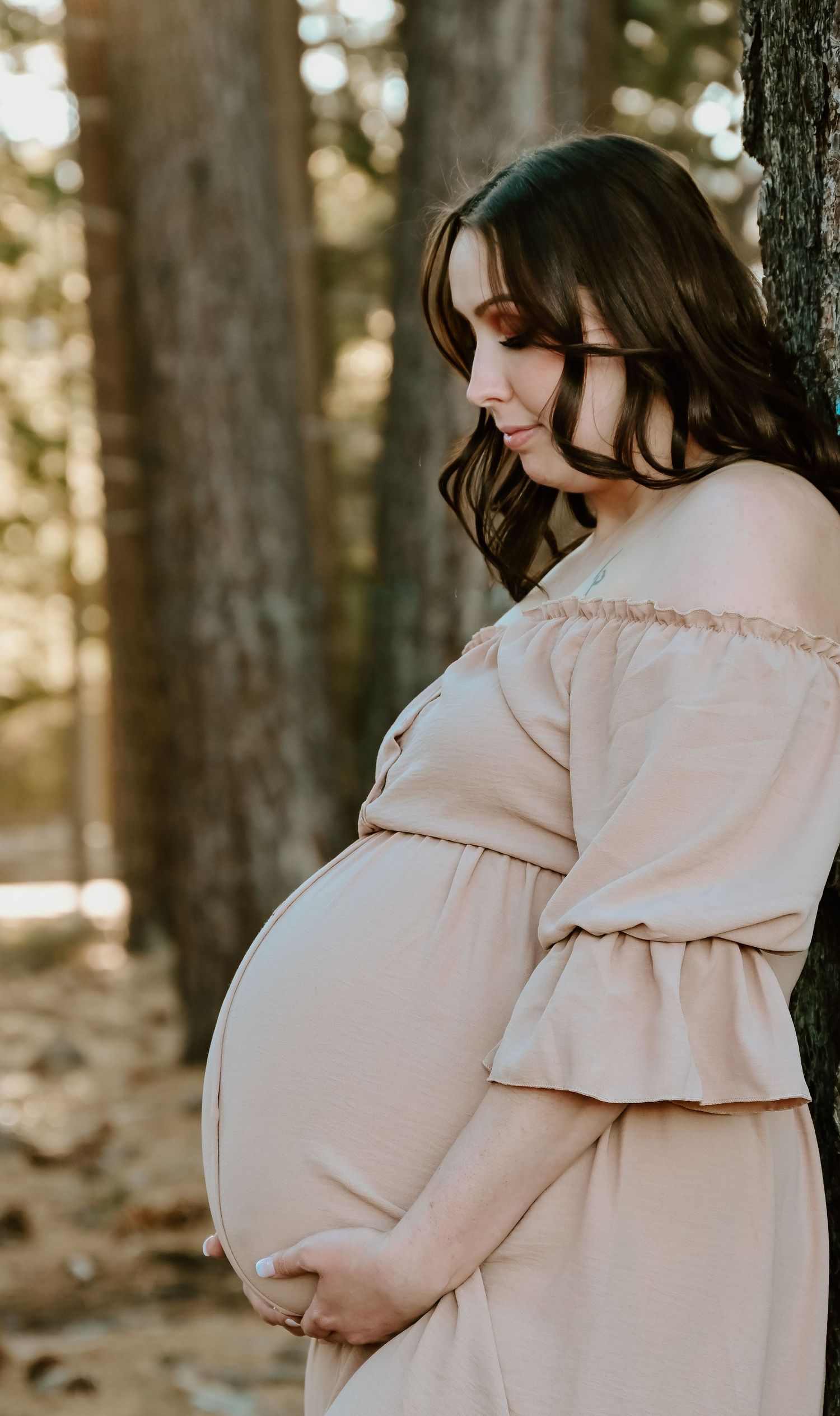 A pregnant person in a soft pink off-shoulder dress stands in a sunlit forest setting, tenderly resting against a tree.