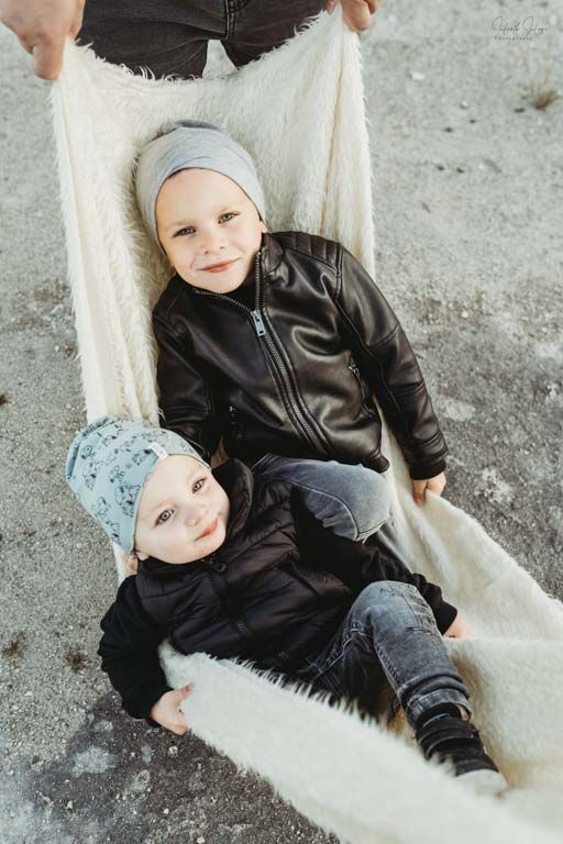 Two children in winter beanies and leather jackets sit together on a white furry blanket outdoors.