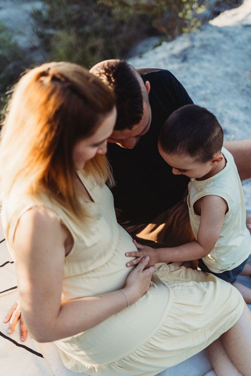 Three people sit together outdoors while looking down at a pregnant woman's belly in a tender family moment.