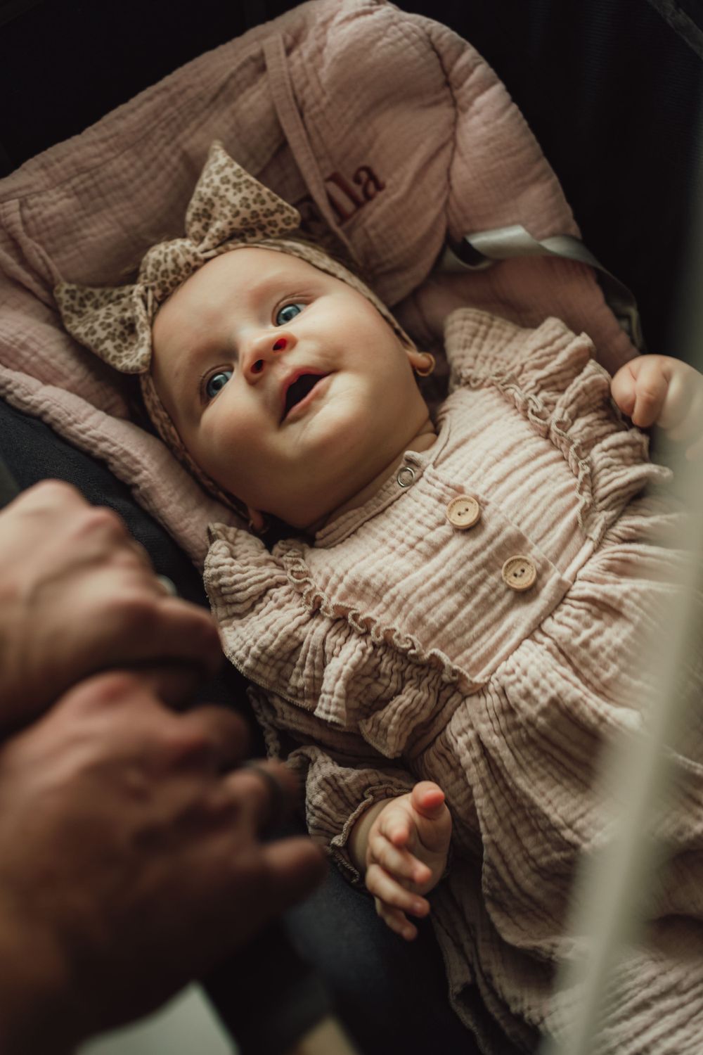 A small baby in a beige ruffled dress and sparkly bow headband smiles while lying down.