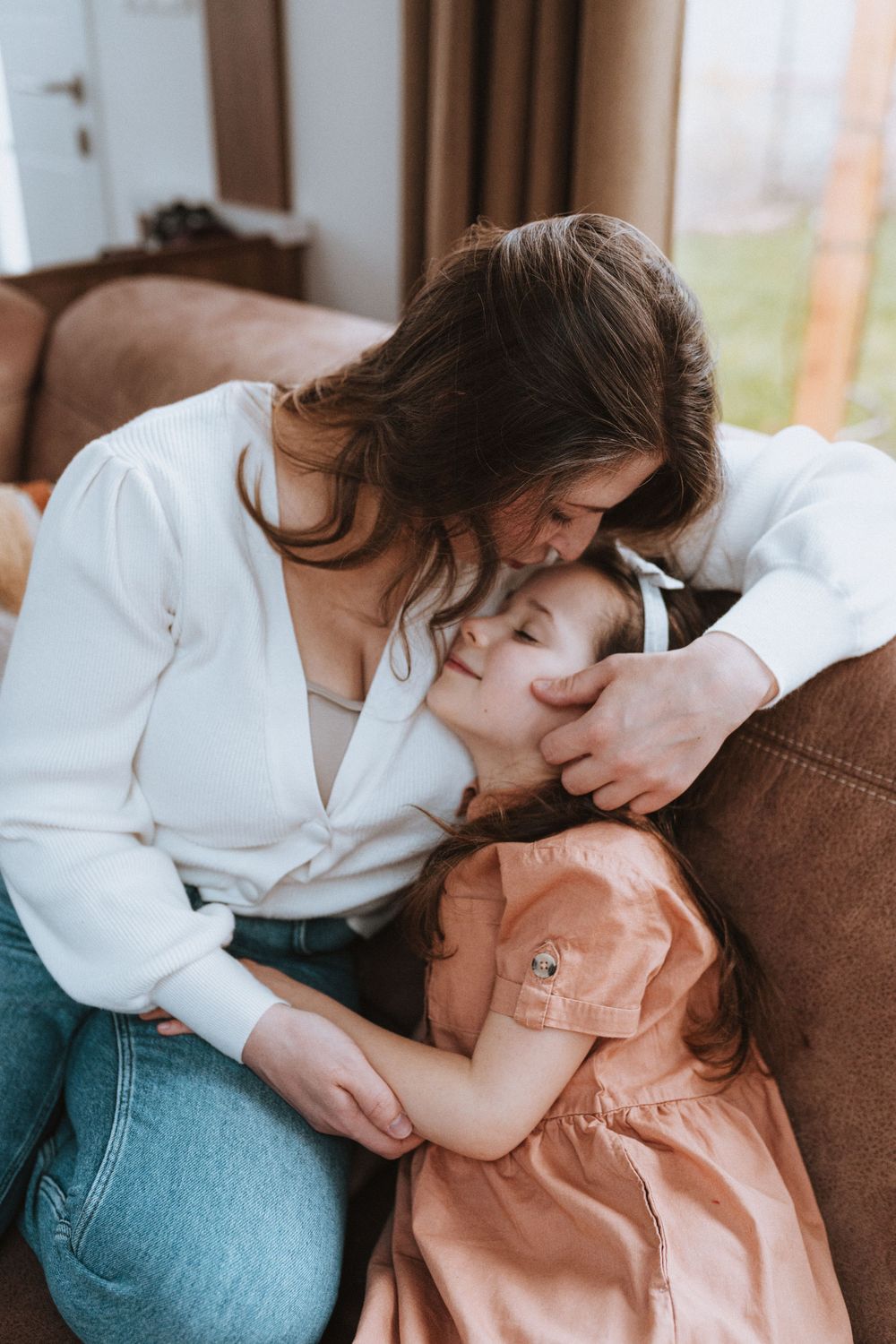 A tender moment shared between a woman in white shirt and jeans embracing a child in a peach dress on a brown couch.