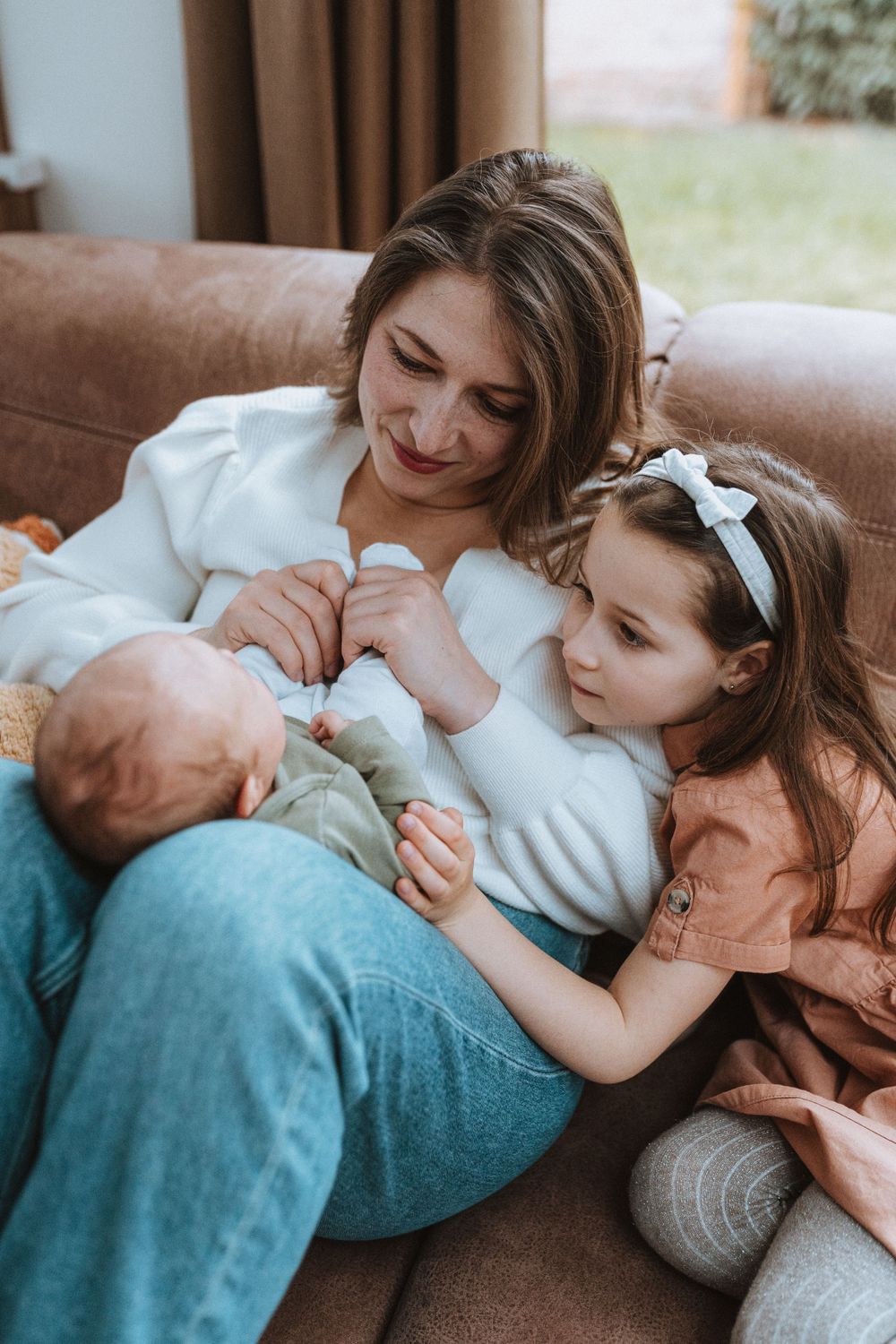A warm family moment on a brown couch as a toddler and parent share a tender moment with a newborn baby.