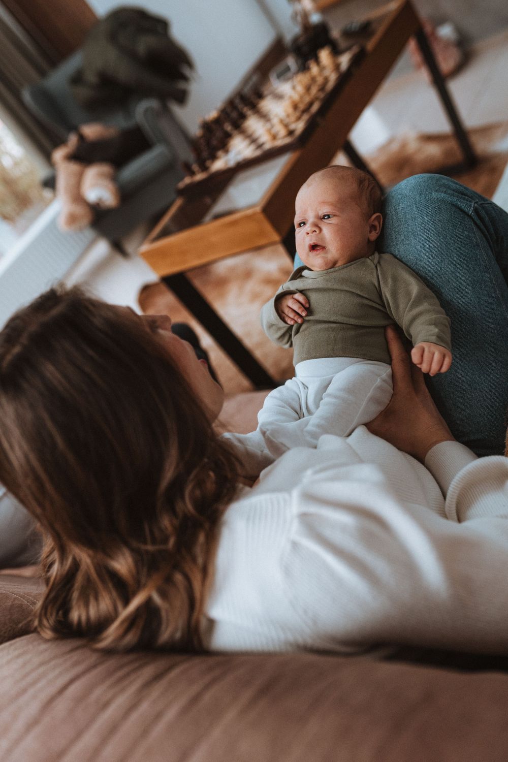 A baby in a green outfit lays on a white blanket while being comforted on a brown couch.