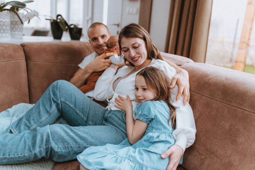 A family relaxes together on a brown couch at home, enjoying a cozy moment in casual clothing.